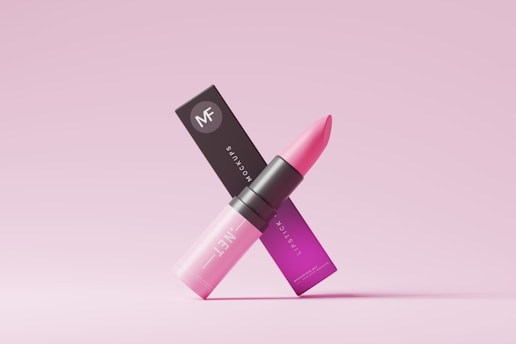 A pink lipstick is positioned in an elegant manner along with its packaging. The lipstick tube is sleek and modern, with a gradient of pink shades transitioning from light at the bottom to a darker hue at the top. The packaging displays the brand logo along with text in a matching pink color scheme. The overall aesthetic is minimalist and chic, set against a light pink background.