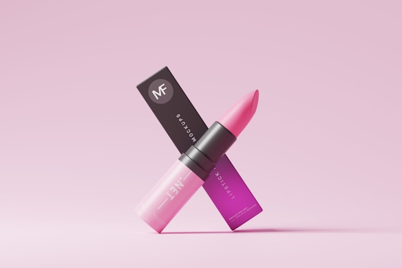 A pink lipstick is positioned in an elegant manner along with its packaging. The lipstick tube is sleek and modern, with a gradient of pink shades transitioning from light at the bottom to a darker hue at the top. The packaging displays the brand logo along with text in a matching pink color scheme. The overall aesthetic is minimalist and chic, set against a light pink background.