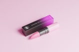 A sleek pink lipstick lies next to its rectangular black and pink box on a matching pink surface. The lipstick is partially opened, revealing its vibrant pink color, and the box is elegantly designed with minimalist typography.