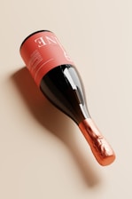 A bottle of wine is placed on its side with a sleek, modern design. The label is a soft pink with white text, while the neck is wrapped in a shiny copper foil. The glass of the bottle is dark, creating a reflective surface that enhances its elegant appearance.
