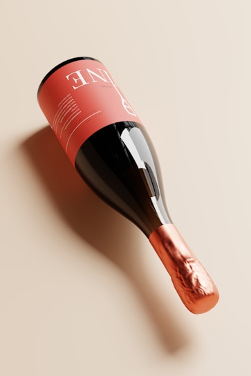 A bottle of wine is placed on its side with a sleek, modern design. The label is a soft pink with white text, while the neck is wrapped in a shiny copper foil. The glass of the bottle is dark, creating a reflective surface that enhances its elegant appearance.