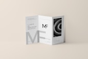 A tri-fold DL brochure mockup on a beige background. The brochure features modern typography and geometric design elements, with one panel displaying a black-and-white image of a spiral pattern. The brochure text includes a prominent 'MF' logo.