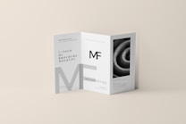 A tri-fold DL brochure mockup on a beige background. The brochure features modern typography and geometric design elements, with one panel displaying a black-and-white image of a spiral pattern. The brochure text includes a prominent 'MF' logo.