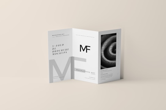 A tri-fold DL brochure mockup on a beige background. The brochure features modern typography and geometric design elements, with one panel displaying a black-and-white image of a spiral pattern. The brochure text includes a prominent 'MF' logo.