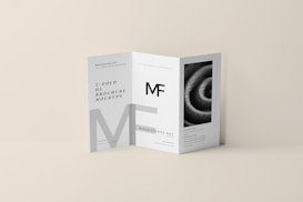 A tri-fold DL brochure mockup on a beige background. The brochure features modern typography and geometric design elements, with one panel displaying a black-and-white image of a spiral pattern. The brochure text includes a prominent 'MF' logo.