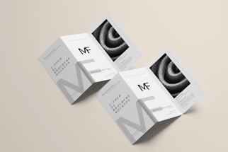 Two unfolded tri-fold brochures are displayed on a light beige background. The design features monochrome elements with the letters 'MF' prominently displayed. The brochures include detailed text and an image of a coiled rope or similar texture.