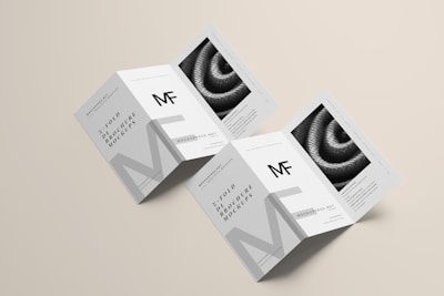 Two unfolded tri-fold brochures are displayed on a light beige background. The design features monochrome elements with the letters 'MF' prominently displayed. The brochures include detailed text and an image of a coiled rope or similar texture.
