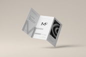 A tri-fold brochure mockup is depicted with a minimalist design. It features clean lines and a modern aesthetic with a grayscale color scheme. The front side has an 'MF' logo, and there's an abstract spiral image on one of the panels.