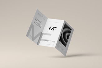 A tri-fold brochure mockup is depicted with a minimalist design. It features clean lines and a modern aesthetic with a grayscale color scheme. The front side has an 'MF' logo, and there's an abstract spiral image on one of the panels.