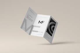 A tri-fold brochure mockup is depicted with a minimalist design. It features clean lines and a modern aesthetic with a grayscale color scheme. The front side has an 'MF' logo, and there's an abstract spiral image on one of the panels.