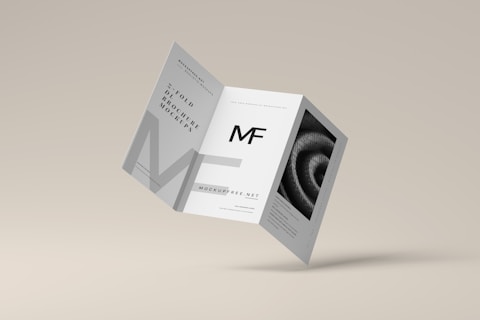 A tri-fold brochure mockup is depicted with a minimalist design. It features clean lines and a modern aesthetic with a grayscale color scheme. The front side has an 'MF' logo, and there's an abstract spiral image on one of the panels.