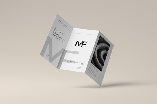 A tri-fold brochure mockup is depicted with a minimalist design. It features clean lines and a modern aesthetic with a grayscale color scheme. The front side has an 'MF' logo, and there's an abstract spiral image on one of the panels.