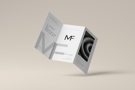 A tri-fold brochure mockup is depicted with a minimalist design. It features clean lines and a modern aesthetic with a grayscale color scheme. The front side has an 'MF' logo, and there's an abstract spiral image on one of the panels.
