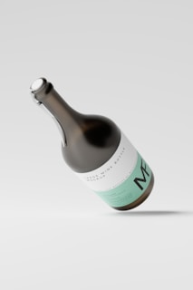 A large wine bottle mockup with a sleek design, featuring minimalistic white and mint green labeling. The bottle is tilted at an angle against a light gray background, casting a soft shadow on the surface.