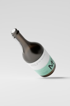 A large wine bottle mockup with a sleek design, featuring minimalistic white and mint green labeling. The bottle is tilted at an angle against a light gray background, casting a soft shadow on the surface.