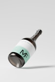 A sleek wine bottle featuring a minimalist design, with a mint green and white label, prominently displaying the initials 'MF'. The bottle is positioned at an angle against a plain, light background, casting a subtle shadow.