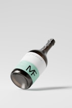 A sleek wine bottle featuring a minimalist design, with a mint green and white label, prominently displaying the initials 'MF'. The bottle is positioned at an angle against a plain, light background, casting a subtle shadow.