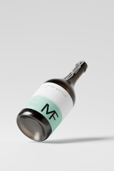 A sleek wine bottle featuring a minimalist design, with a mint green and white label, prominently displaying the initials 'MF'. The bottle is positioned at an angle against a plain, light background, casting a subtle shadow.