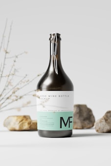 A large wine bottle featuring a modern label design stands in the foreground. The label is white with minimalist text and teal accents that include the letters 'MF'. Behind the bottle, there are some natural elements such as rocks and branches, providing an organic and simple backdrop. The light gray background complements the bottle's dark glass.