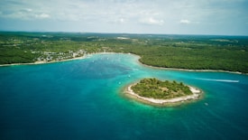 An aerial view of a small, forested island surrounded by turquoise waters, set against a backdrop of a larger, verdant landmass. Numerous boats are scattered in the water, and a few white wakes from moving boats are visible.