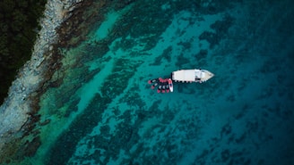 An aerial view of a serene sea with clear turquoise water and a rocky coastline. A white boat is anchored near the shore surrounded by a group of kayaks or small boats.