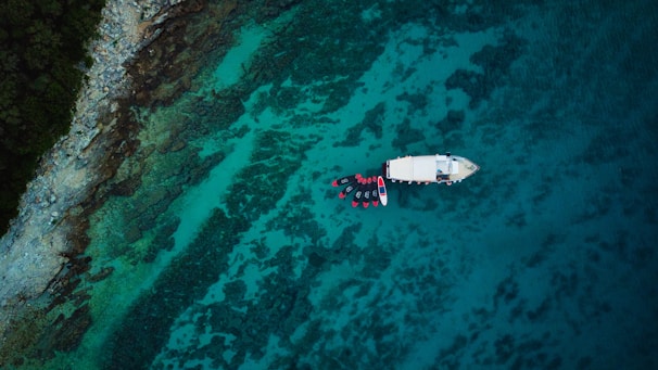 An aerial view of a serene sea with clear turquoise water and a rocky coastline. A white boat is anchored near the shore surrounded by a group of kayaks or small boats.