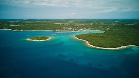 Aerial view of a coastal landscape with lush green forests, a small island, and turquoise waters. The shoreline is dotted with boats, and a small settlement can be seen in the background.