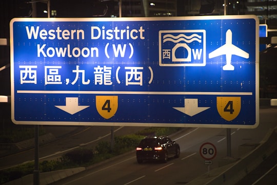 A large blue road sign directing traffic towards the Western District and Kowloon West, featuring white text both in English and Chinese. The sign includes icons indicating airport access and tunnel passage. Below the sign, there's a dark car driving on an illuminated road.