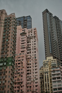 Several high-rise residential buildings are tightly clustered together against a backdrop of overcast skies. The architecture features a mix of modern and older styles with a variety of colors, from pastel pinks and beiges to grey and blue hues. Numerous windows and balconies are visible, giving an urban, densely populated feel.