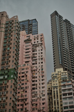 Several high-rise residential buildings are tightly clustered together against a backdrop of overcast skies. The architecture features a mix of modern and older styles with a variety of colors, from pastel pinks and beiges to grey and blue hues. Numerous windows and balconies are visible, giving an urban, densely populated feel.