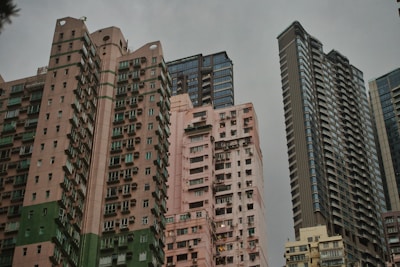 A cluster of high-rise residential buildings with varying architectural styles and colors, set against a gray, overcast sky. The buildings are closely packed, showcasing a mixture of modern and older structures, with visible air conditioning units and windows.