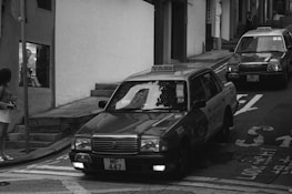 Two taxis driving up a narrow, inclined urban street with a person standing on the sidewalk and another person visible in a shop window. The street is lined with buildings and a traffic sign is partially visible.