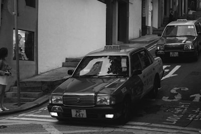 Two taxis driving up a narrow, inclined urban street with a person standing on the sidewalk and another person visible in a shop window. The street is lined with buildings and a traffic sign is partially visible.