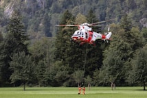 A red and white rescue helicopter hovers above a grassy field, surrounded by dense green forest. Two rescue personnel in orange suits are suspended below the helicopter, connected by a line, engaged in a lifting operation.