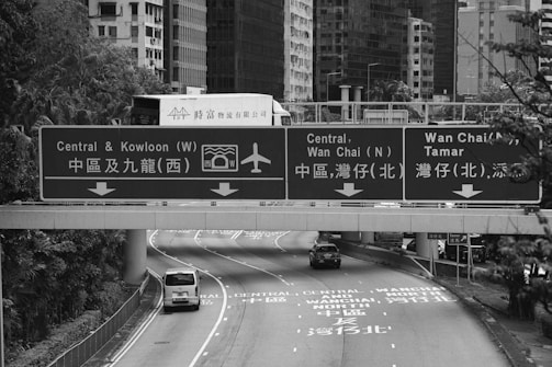 Overhead highway signs guide drivers towards Central, Kowloon, and Wan Chai. Vehicles, including a van and a car, travel on the road beneath. The background features tall buildings and greenery.