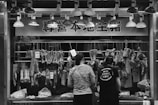 At a butcher's stall, various cuts of meat are displayed hanging on hooks. Two people are standing in front, likely making a purchase. The area is illuminated by a row of overhead lights. Prices are visible on some meat pieces, written in a local currency. The scene is in black and white, adding a timeless, documentary feel.