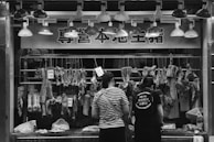 At a butcher's stall, various cuts of meat are displayed hanging on hooks. Two people are standing in front, likely making a purchase. The area is illuminated by a row of overhead lights. Prices are visible on some meat pieces, written in a local currency. The scene is in black and white, adding a timeless, documentary feel.