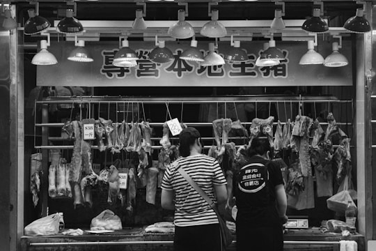 At a butcher's stall, various cuts of meat are displayed hanging on hooks. Two people are standing in front, likely making a purchase. The area is illuminated by a row of overhead lights. Prices are visible on some meat pieces, written in a local currency. The scene is in black and white, adding a timeless, documentary feel.