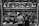 At a butcher's stall, various cuts of meat are displayed hanging on hooks. Two people are standing in front, likely making a purchase. The area is illuminated by a row of overhead lights. Prices are visible on some meat pieces, written in a local currency. The scene is in black and white, adding a timeless, documentary feel.