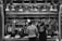 At a butcher's stall, various cuts of meat are displayed hanging on hooks. Two people are standing in front, likely making a purchase. The area is illuminated by a row of overhead lights. Prices are visible on some meat pieces, written in a local currency. The scene is in black and white, adding a timeless, documentary feel.
