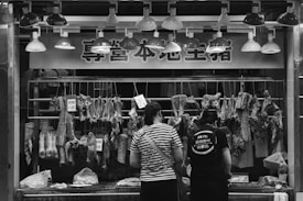 At a butcher's stall, various cuts of meat are displayed hanging on hooks. Two people are standing in front, likely making a purchase. The area is illuminated by a row of overhead lights. Prices are visible on some meat pieces, written in a local currency. The scene is in black and white, adding a timeless, documentary feel.