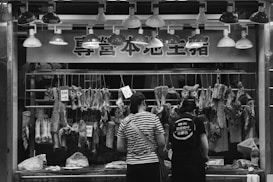 At a butcher's stall, various cuts of meat are displayed hanging on hooks. Two people are standing in front, likely making a purchase. The area is illuminated by a row of overhead lights. Prices are visible on some meat pieces, written in a local currency. The scene is in black and white, adding a timeless, documentary feel.