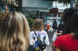 A group of people walk along a city street, with some in focus wearing colorful attire. One person is wearing a shirt with 'New Orleans' written on the back. The street is lined with shops, and there are blurred images of storefronts and signs in the background.