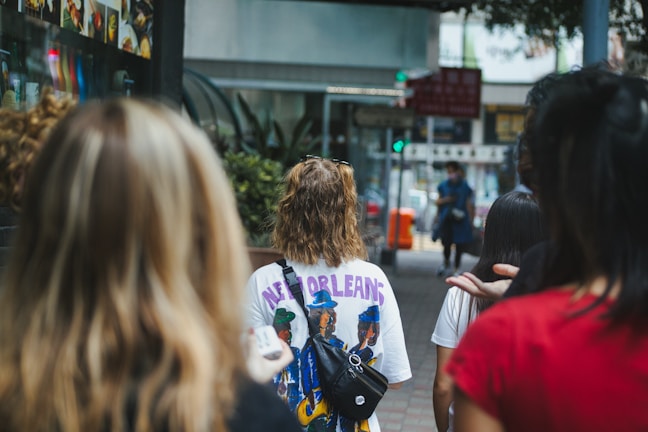 A group of people walk along a city street, with some in focus wearing colorful attire. One person is wearing a shirt with 'New Orleans' written on the back. The street is lined with shops, and there are blurred images of storefronts and signs in the background.
