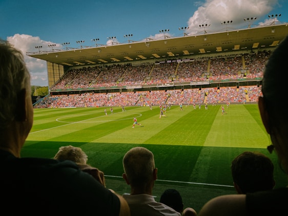 A large soccer stadium filled with spectators and two teams playing on a lush green field. The stands are packed with fans wearing colorful clothing, and the stadium roof is visible under a bright blue sky with scattered clouds.