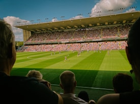 A large soccer stadium filled with spectators and two teams playing on a lush green field. The stands are packed with fans wearing colorful clothing, and the stadium roof is visible under a bright blue sky with scattered clouds.