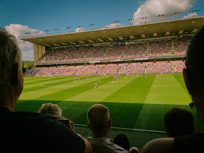 A large soccer stadium filled with spectators and two teams playing on a lush green field. The stands are packed with fans wearing colorful clothing, and the stadium roof is visible under a bright blue sky with scattered clouds.