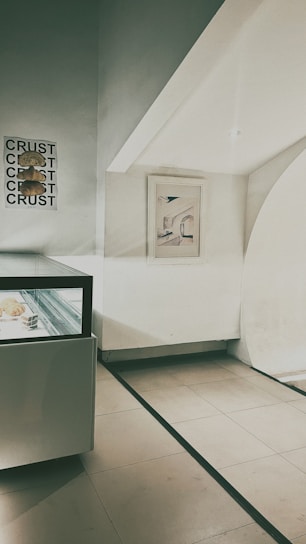 A minimalistic interior scene with a pastry display case containing baked goods, a framed abstract artwork on the wall, and a poster displaying the word 'CRUST' multiple times alongside images of bread.