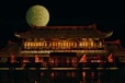 A traditional Vietnamese building illuminated at night with an intricate roof and multiple windows, adorned with red and gold lights. A large patterned circle with text in Vietnamese is displayed in the background, and several red and yellow flags are situated in front of the building.
