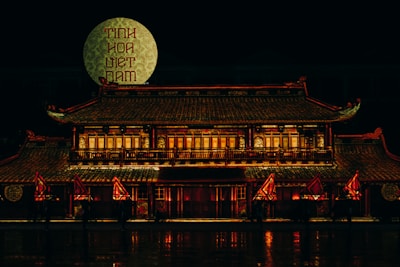A traditional Vietnamese building illuminated at night with an intricate roof and multiple windows, adorned with red and gold lights. A large patterned circle with text in Vietnamese is displayed in the background, and several red and yellow flags are situated in front of the building.